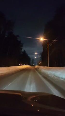 The video shows a driver's perspective of a car moving along a snowy road at night. The road is lined with tall, dark trees on either side, and there are streetlights illuminating the road ahead. Patches of snow cover the shoulders of the road. The sky is dark, with a few clouds visible. The camera is static, fixed inside the car, and there are no other discernible actions or interactions in the frame.