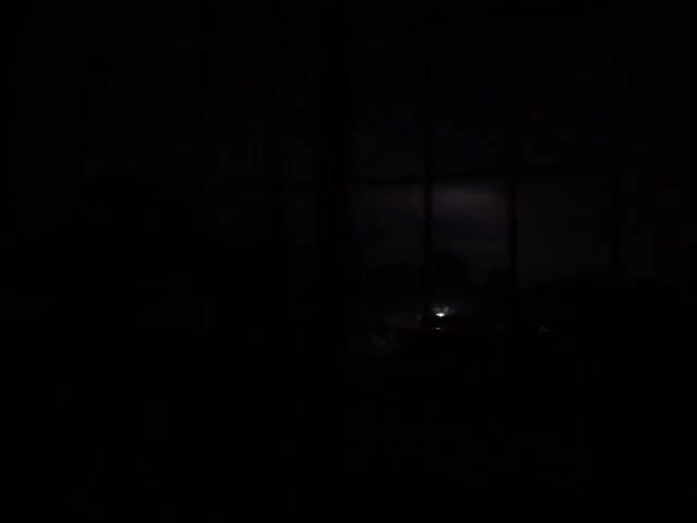 The video shows a view from inside a room through a window with a grid pattern. Outside, it is dark, and there is a bright flash of lightning. The light briefly illuminates a blurry scene, possibly showing trees or buildings in the distance.
