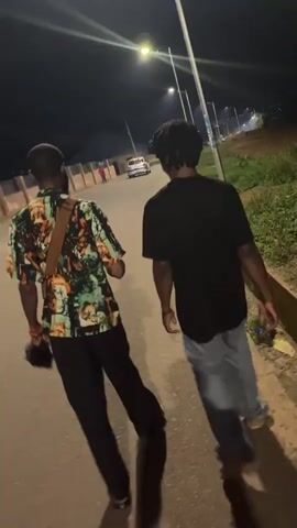 Two individuals, seen from behind, walk down a paved road at night. The person on the left wears a short-sleeved shirt with a colorful, abstract pattern and black pants. They carry a black bag in their left hand and a black object, possibly a hat, is attached to their right wrist with a red string. A tan strap crosses their chest from their right shoulder. The person on the right wears a black t-shirt and light blue pants. Both individuals cast long shadows on the road, illuminated by a street light visible in the upper portion of the frame. The road is bordered on the right by a raised concrete curb and grassy area. In the background, a few structures and a car are faintly visible under the night sky. The overall scene suggests a casual nighttime walk on a street.