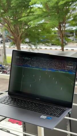 A laptop displays a soccer match on its screen, with players on a green field and spectators in the background. The laptop is placed on a balcony railing, with lush green trees and a street visible behind it. Cars and motorcycles are parked on the street, and some buildings are in the distance. The sky appears to be overcast.