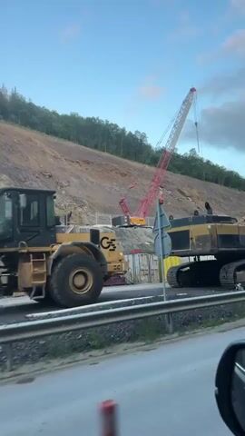 The video shows a large construction site alongside a road, featuring several pieces of heavy machinery and construction materials. In the foreground, a large yellow tracked excavator, possibly a Caterpillar given the visible "CAT" logo, is parked next to a guardrail.

Behind the excavator, a large red and white lattice boom crane, potentially with "HK" visible on its boom, stands tall against a steep hillside. The hillside, partially covered with trees at the top, shows areas of exposed earth and rock, suggesting excavation work.

Further along the site, various construction elements are visible: large concrete pipes and smaller metal pipes are laid out on the ground. Several small white portable container offices or sheds are present, along with a blue portable toilet. Another red excavator with a hydraulic hammer attachment is positioned on the slope, and a yellow backhoe loader and a road roller are also seen.

The scene suggests ongoing or recently completed infrastructure development, possibly a road expansion or stabilization project, given the hillside and pipes. The sky is blue with scattered clouds, indicating fair weather. The soft light suggests it's late afternoon or early evening. The video is filmed from a moving vehicle, likely a car, passing by the construction area.
