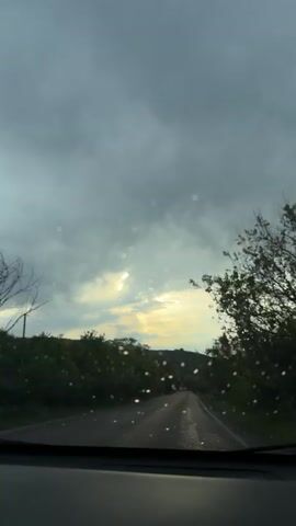 The video shows a view from inside a car driving on a wet asphalt road under a cloudy sky. Sunlight peeks through a gap in the clouds, creating a bright spot on the horizon. Trees line the sides of the road, and a distant hill is visible. Other cars are driving on the road ahead. There are raindrops on the windshield, and music is playing in the background.