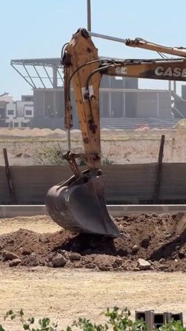 An excavator is digging a hole in the ground. The excavator is yellow and orange and has the brand name CASE on it. A person is inside the cab of the excavator, operating it. The ground is dirt and there are some rocks and debris. In the background, there are buildings under construction and a blue sky. The video is taken on a sunny day.
