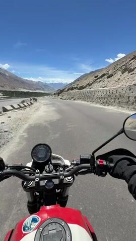 The video begins with a first-person perspective of someone riding a red and white motorcycle on a paved road. The rider's hands are on the handlebars, and a circular digital display is visible on the dashboard. The landscape is arid and mountainous, with rocky slopes on either side of the road. The sky is a clear blue with a few scattered white clouds. As the motorcycle moves, the scenery shifts, revealing more of the vast, barren terrain and distant mountain ranges. A mirror attached to the handlebar reflects the rider's helmet and the surrounding environment. The road appears to be in a remote, possibly high-altitude region, given the rugged topography and lack of vegetation. The video continues to show the motorcycle driving along the road, with the camera capturing the expansive views of the mountains and valleys. The sun is bright, indicating daytime. There are no other vehicles or people visible in the immediate vicinity of the motorcycle.