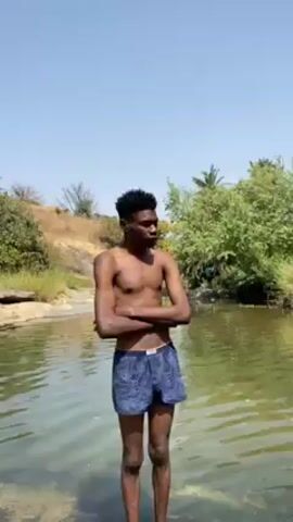 A young, slender, shirtless Black man with short curly hair stands in water. He wears blue boxer shorts with a floral pattern. He is standing in a body of water, surrounded by green trees and a grassy bank. The sky is clear and blue. The man crosses his arms, smiles, and then uncrosses them. The water appears calm.