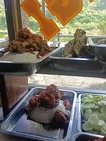 The video pans across a food stall, showcasing various dishes in metal trays. In the foreground, a tray contains what appears to be rice topped with a reddish sauce and meat. To its right, a tray is filled with sliced cucumbers. Behind these, in the upper section, several more trays hold different food items. One contains a mound of reddish-brown food, possibly meat in sauce. Another tray has a pile of fried items, perhaps chicken or fish. A third tray holds a generous portion of a yellowish, granular food. Further back, more trays display what look like dried ingredients or snacks, some dark brown and others lighter brown, resembling small fish or fried vegetables. The background shows a window with a view of a green, hilly landscape, suggesting an outdoor or semi-outdoor setting. The lighting appears to be natural daylight. There are no people clearly visible in the frame, and no specific actions or interactions are captured. The focus is on the food displays.