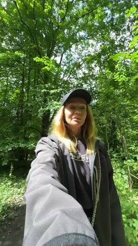 A woman wearing a black baseball cap, a dark jacket over a black t-shirt, and a necklace with a chain walks through a sun-dappled forest. She is holding a camera in her right hand, which is pointing forward, suggesting she is filming herself. The forest is lush with green trees and foliage, and a dirt path runs alongside a body of water. The woman appears to be looking off to the side, possibly at the scenery or at something she's encountering on her walk. The video captures the natural beauty of the forest environment.