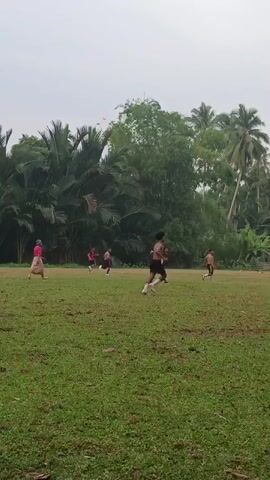 This video shows an outdoor soccer game played on a grass field. Several young men are on the field, some wearing shorts and athletic tops, others shirtless. The game appears to be informal, with no clearly defined teams or spectators. The field is surrounded by lush green vegetation, including palm trees and other tropical plants, suggesting a warm climate. The sky is overcast, with no visible sun. The camera is handheld and pans across the field, following the action of the game. There is no commentary or dialogue. The overall impression is one of a casual, community-based sporting event taking place in a natural, tropical setting.