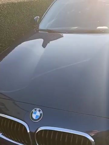 The camera slowly pans upward and to the right, revealing a navy blue BMW hatchback parked on a gravel driveway. The car is facing the camera and is positioned in front of a short, green, trimmed hedge. The sun glares off the car's hood and windshield, creating a bright reflection. The car appears clean and well-maintained. The license plate is partially visible. The environment appears to be a residential driveway.