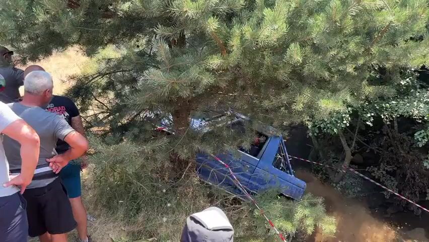 A group of men stand around a pine tree, some looking at a blue vehicle that is off-road and partially obscured by branches. The vehicle appears to be stuck in a ditch or muddy area, with its wheels on the side. Red and white tape is visible in the foreground, possibly marking a boundary or safety zone. The environment is outdoors with dry grass and other trees in the background.