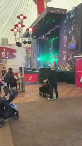 The video shows a man standing behind a stroller, facing a stage where two performers in red costumes are juggling hula hoops. A young girl in a white jacket and patterned pants walks toward the man and stroller. The stage is decorated with musical notes and has green and red stage lights.  The area appears to be an indoor public space with a patterned tile floor and decorative lamp posts. There are other people in the background and additional strollers.