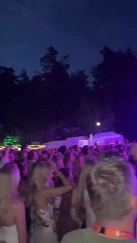 The video shows a concert with a live performer on a stage under bright purple lights. The audience is visible in the foreground, with many people holding up their phones to record. The performer, wearing a white t-shirt and blue shorts, is holding a microphone and gesturing with his free hand. The stage is set up with speakers and lighting equipment. The atmosphere appears energetic and festive.