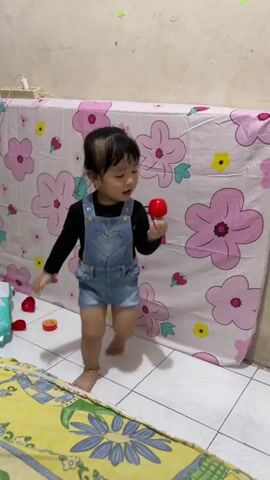 A toddler girl, wearing a black long-sleeved shirt and denim overalls, is being fed by someone's hand holding a spoon with food. The girl is holding a red toy in her right hand and has a bowl with some food in front of her. The background shows a wall and a bed with a pink floral pattern. The floor is tiled and there is a yellow blanket with a blue floral pattern in the foreground.