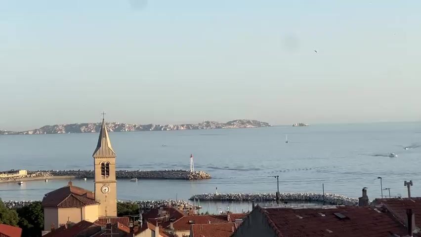 A long shot shows a coastal town with buildings and a church with a steeple in the foreground. Beyond the town is a harbor with two breakwaters, one with a lighthouse. Small boats are visible in the harbor and in the open sea. In the distance, a chain of islands stretches across the horizon under a clear sky. The sun appears to be low in the sky, casting a warm light on the scene.