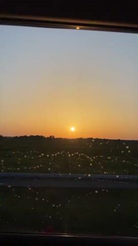 The video shows a sunset viewed from inside a vehicle, possibly a car. The sun is a bright orb low on the horizon, casting an orange and yellow glow across the sky. In the foreground, out of focus, are water droplets on the car window, creating circular blurs of light. Beyond the window is a dark silhouette of a line of trees or hills, and further beyond that, a field with scattered white dots, possibly flowers or other vegetation. A dark metal railing is visible in the lower portion of the frame, suggesting the vehicle is on a road or bridge. The overall scene conveys a sense of peaceful travel during sunset.