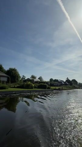 The camera moves slowly across a smooth body of water. On the left and right are houses with green lawns and trees going down to the water's edge. The houses are reflected in the water. The sky is blue with a few wispy clouds.