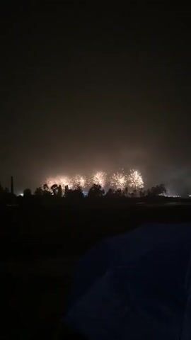 The video shows a nighttime scene with fireworks exploding in the distance. The sky is filled with a bright red glow from the fireworks and smoke. In the foreground, a dark blue umbrella is visible, partially obscuring the bottom of the frame. Trees and buildings can be seen silhouetted against the illuminated sky. The ground appears wet, possibly from rain. The fireworks are varied, with some creating large bursts of light and others shooting upwards in columns. The overall atmosphere is one of a celebratory event.