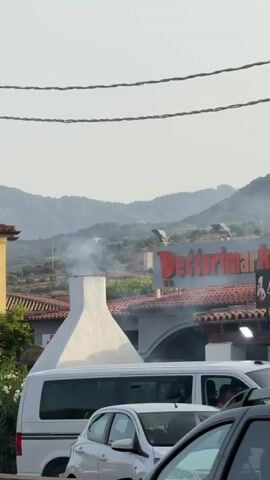 This is a vertical video with a slightly hazy sky. In the foreground, the tops of cars are visible, indicating a traffic situation. In the middle ground, a large white chimney with smoke emanating from its top is prominent. Behind the chimney, a building with a red sign that reads "Dettorimarket" is visible, along with red-tiled roofs. Mountains with sparse vegetation form the background. A person is partially visible near the base of the chimney. The overall impression is an outdoor scene, possibly in a rural or semi-rural commercial area, with some ongoing activity suggested by the smoke.