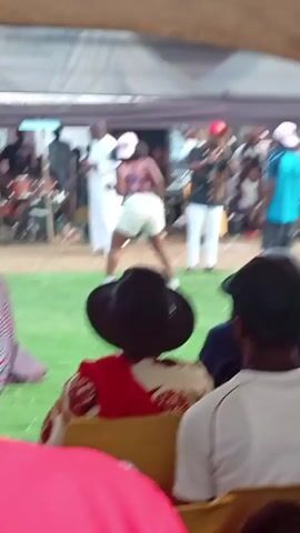 The video shows an outdoor event under a tent, with people watching a female dancer perform on a grassy area. The dancer is wearing shorts, a top, a white cap, and white sneakers. Behind her, a band is playing music, and a man with a microphone is addressing the crowd. The audience is seated on chairs under the tent, observing the performance. There is a table with beverages and cups nearby. The overall setting suggests a celebratory or festive occasion.