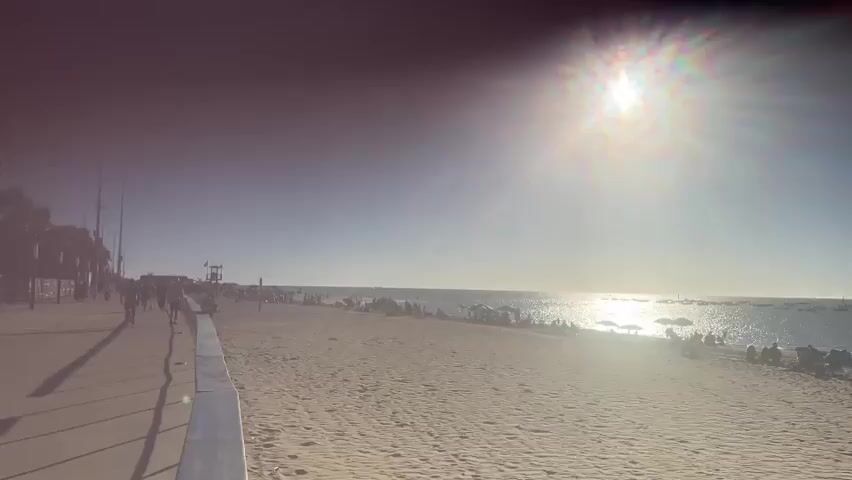 A wide shot of a beach shows a sandy shore with people scattered across it, some sitting on lounge chairs under umbrellas. The ocean stretches out to the horizon, dotted with several boats. The sun is high in the sky, casting a bright glare on the water and creating lens flare effects in the footage. On the left side of the frame, a paved walkway runs parallel to the beach, with people strolling along it. The overall scene depicts a sunny day at a popular beach destination.