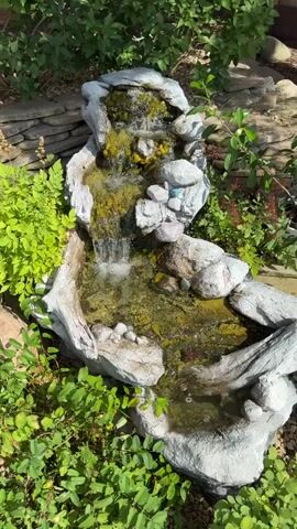 A close-up shot of a multi-tiered artificial waterfall in an outdoor garden. The waterfall is constructed with gray, rock-like material and features yellow algae growth on its surfaces. Water cascades down from the top level, creating a small pool at the bottom. The surrounding area is filled with lush green foliage and a stone wall in the background. The video captures the gentle flow of water and the natural setting.