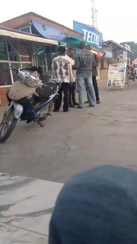 The video shows a small group of men standing in a street. A motorcycle is parked to the left. The men appear to be talking or looking at something in one of their hands. Behind them are small shops, one with a "Tecno" sign. A car passes by in the background. The video is shot from a low angle.