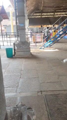 A video shows a large, open-air covered structure, possibly a temple or market. The floor is made of large stone tiles, some of which are wet and reflective, indicating recent cleaning or rain. In the foreground, a large stone pillar is visible on the left. To the right of the pillar, a green trash can is present. Further back, a blue metal staircase leads upwards. Several people are visible in the background, some walking and some standing. The overall lighting suggests it is daytime. The camera is handheld and moves slightly throughout the recording.