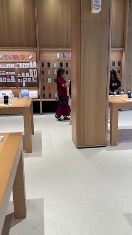 A retail store, possibly an electronics store, features wooden display tables showcasing mobile phones. The walls are lined with shelves displaying various phone accessories and boxed items. Several people are browsing the merchandise. In the foreground, a person wearing a red dress is examining a phone on a display table. In the midground, a man with his hair in a bun and a woman in a red outfit are looking at phones. Further back, other customers are visible, some interacting with staff or products. The overall environment appears clean and well-lit, with a polished floor.
