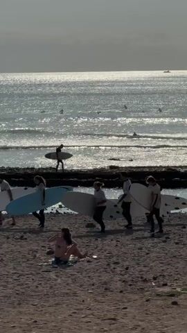 The video captures a beach scene with several people carrying surfboards. In the foreground, a person is sitting on the sand, facing away from the camera. Behind them, a group of individuals are walking with surfboards, some on the sand and some near the water's edge. Further out in the ocean, more people on surfboards can be seen in the water. The sun is setting, casting a glint on the water. The environment appears to be a rocky beach with the ocean stretching to the horizon.