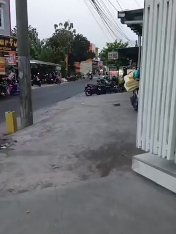 A street scene in what appears to be a Southeast Asian country. The time of day seems to be late afternoon or early evening, as the lighting is subdued. The street is paved and lined with buildings and trees. Several motorbikes are parked along the side of the road, and a few are in motion. People are visible on the motorbikes and walking on the sidewalk. There are signs in a foreign language on the buildings. A power pole with numerous wires is visible in the foreground.