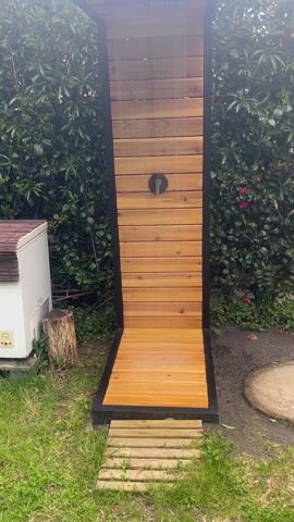 The video shows an outdoor shower made of wood and black metal. The showerhead is square and attached to the top of the structure, and water is flowing from it. The shower has a wooden floor with a small wooden pathway leading up to it. The shower structure is surrounded by green bushes and grass. Next to the shower is a small white appliance or cooler and a wooden stump. The camera pans up and back, showing the full structure of the outdoor shower.