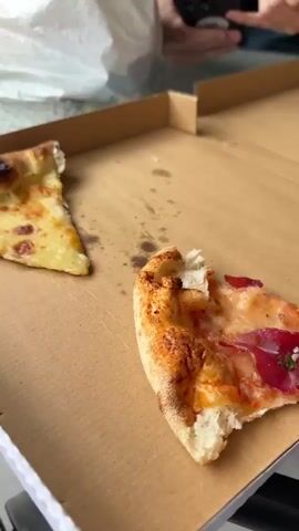 The video shows a table on a patio with a slice of pizza in a box and a protein bar on it.  The patio is wet from rain.  A decorative white block wall runs along the edge of the patio.  Buildings are visible in the background.