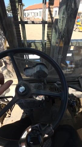 The video shows the interior of a forklift from the driver's perspective. The steering wheel is large and black, with a dusty control panel below. The forklift mast and chains are visible through the dirty windshield, framing a view of a sunny outdoor area. Several buildings with white walls and red-tiled roofs are visible in the background. A yellow forklift is also visible, parked further away. The driver's left hand is briefly visible on the steering wheel. The scene seems to be taking place in a work yard or construction site.