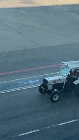A white airport ground support tractor with a "wfs" logo and "ORG-1834 LMO1834" identifier on its roof canopy drives from left to right across an asphalt tarmac. A person in a plaid shirt is driving the vehicle, which has a yellow and black striped bumper and a tow hitch. Other ground support vehicles, including a white Renault Duster car and larger tugs, are parked in the background. Pink and yellow markings are visible on the tarmac.