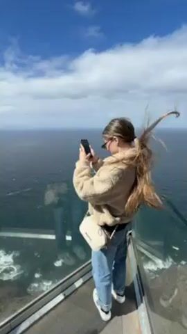 The video shows a woman standing on a glass observation deck, taking a photo with her phone. She has brown hair pulled back into a ponytail and is wearing a beige sherpa jacket, jeans, and white sneakers. The deck overlooks a rugged coastline with steep, dark cliffs meeting the ocean. The sea is choppy with whitecaps visible where the waves crash against the rocks. Above, the sky is bright blue with scattered white clouds, and the sun is shining. The glass panels of the deck offer a clear view of the water and the landscape.