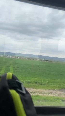 The video shows a view from inside a vehicle, possibly a bus, looking out a window at a green field stretching to the horizon under a cloudy sky. The field appears to be crops, with a farm or small settlement visible in the distance. A dirt road runs alongside the field. The foreground includes a portion of a backpack, black with a neon yellow trim, resting on the window frame or a nearby seat. There are no visible people or actions other than the movement of the vehicle. The scene is dominated by the landscape and weather conditions.