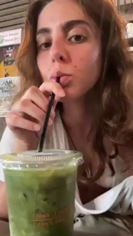 A young woman with brown hair and fair skin is drinking from a plastic cup filled with a green, iced beverage, using a black straw. She is wearing a white top with a lace-edged camisole underneath. She is sitting at a wooden table. In the background, there is a wall decorated with signs and posters. Her necklace is visible. The video appears to be shot indoors.