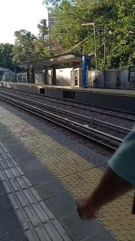 The video shows a train station platform. The camera pans across the platform and the train tracks. A person with a bicycle is standing on the platform. There are other people on the platform, but they are not the focus of the video. The platform is marked with yellow tactile paving. The sky is visible above the trees in the background.