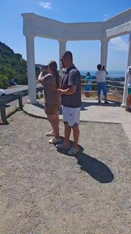 The video pans across an outdoor scenic overlook on a sunny day. Several people, dressed in casual summer clothing, are scattered around a white, open-air structure supported by columns. Some individuals are taking photographs with their phones, while others are simply observing the view. In the foreground, two people are standing side-by-side, one holding a phone. In the background, a body of water and a distant coastline are visible. The ground is covered in gravel and concrete.
