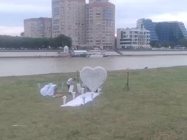 A person is setting up a wedding ceremony area on a grassy bank by a river. The area is decorated with a large white heart made of balloons, a white aisle runner, white vases with flowers, and a small table with a blue tablecloth and white decorations. In the background, there are several large buildings and a boat on the river. The sky is overcast.