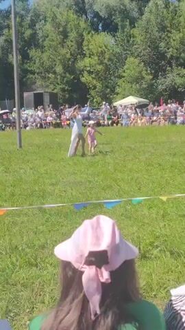 A short video captured from behind a seated spectator shows a grassy field under a sunny sky with a line of trees in the background. A white banner with colorful triangular flags strung across it is visible in the foreground. In the middle ground, two individuals are performing or dancing on the grass in front of an audience seated in rows and standing around. The person in front appears to be an adult in a white outfit, possibly a dress with a long train, and is holding a thin object. A younger child in a pink outfit is positioned slightly behind and to the side of the adult. The audience is spread out, with some people seated on chairs and others standing. Some are holding umbrellas or wearing hats to shield themselves from the sun. Several tents or temporary structures are also visible in the background behind the audience. The overall scene suggests an outdoor event or performance taking place in a park or open field.