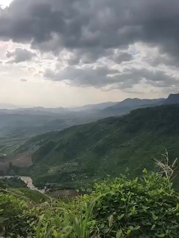 This is a landscape video of rolling green hills and mountains under a cloudy sky. In the foreground, there are some green plants and leaves. A river or stream winds through the valley below. The overall scene is one of natural beauty and vastness. The video appears to be shot from a high vantage point, looking out over the expansive terrain. There are no people or man-made structures prominently featured in the footage. The lighting suggests it may be early morning or late afternoon, with the sun peeking through the clouds.