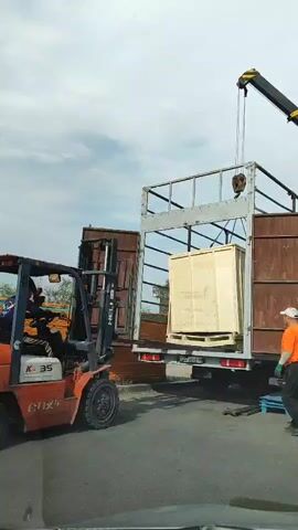 A forklift is lifting a large wooden crate into the back of a trailer. A crane is also attached to the crate, assisting in its movement. A man in an orange shirt is standing next to the trailer, observing the operation. The scene appears to be an industrial or logistical area, possibly a warehouse or loading dock. The sky is overcast, suggesting it might be a cloudy day. The forklift is orange and has "HELJ" and "K-35" visible on its side. The man in the orange shirt has "SENYO" printed on the back of his shirt. The overall atmosphere seems to be one of work and logistics.