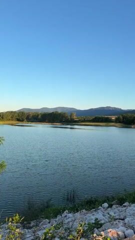 The video opens with a view of a calm body of water under a clear blue sky. The water's surface is gently rippled. The foreground shows a rocky embankment along the edge of the water. As the camera pans to the right, a concrete path emerges, running alongside the embankment and leading away from the viewer. The path is bordered by grass and small stones on one side and the water on the other. Further along the path, the landscape opens up to reveal rolling hills covered in trees and fields, with more distant mountains forming the horizon. The sun is low in the sky, casting warm light and long shadows across the scene. The overall impression is one of a peaceful, natural outdoor setting, possibly a park or a recreational area.