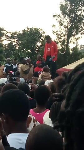 A group of people are gathered outdoors. In the center, a man in a red shirt with white writing on it stands on a raised platform or surface. He appears to be speaking to the crowd. The crowd is mostly facing the man on the platform, with some individuals looking down, possibly in prayer. The lighting suggests it is either early morning or late afternoon. Trees and foliage are visible in the background, indicating a natural or semi-natural environment. The image is slightly blurred and has a vertical orientation.