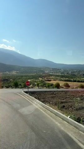 A video shows a landscape with mountains in the distance, a valley with trees and fields, and a road in the foreground. The sky is blue with some clouds. The camera is moving, likely from a vehicle, giving a dynamic view of the scenery. The foreground is a dirt and rocky surface, suggesting the path of movement.