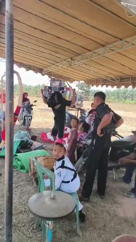 A group of people are gathered under a tan tarp canopy outdoors. In the center, a person with long dark hair is dancing energetically, arms raised. To their right, a child in a pink dress and two other children stand near a motorcycle. A man in a black shirt and pants, possibly a security guard, stands facing the dancer. To the left, a person in a white uniform with a red and white headdress is seated on a chair, appearing to hold drumsticks, with a cymbal in front of them. Several drums and green rolled-up materials are on the ground. In the background, motorcycles and more people are visible, along with trees and an open, possibly rural, environment. The overall atmosphere appears to be a lively outdoor event.