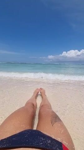 The video shows a person sitting on a white sand beach with their legs extended towards the ocean. The water is clear and turquoise, with small waves lapping at the shore. The sky is a bright blue with a few white clouds. The person is wearing a dark blue and red swimsuit bottom. There is a tattoo on the person's thigh. The camera pans up to show the horizon and the clear sky, then back down to the person's legs.