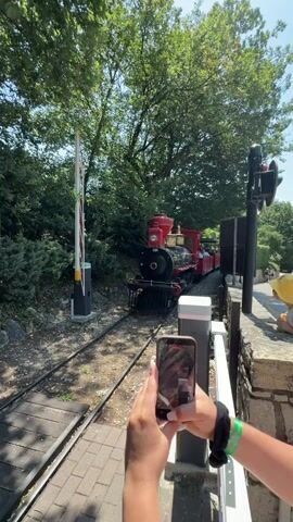 A person's hands hold a smartphone and film a red and black miniature train as it passes by on a sunny day. The train, with passengers in the cars, is on tracks that run alongside a brick walkway and a stone wall. Trees and greenery surround the scene. A railroad crossing gate is visible to the left of the train.