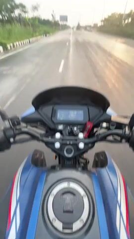 The video captures a first-person perspective of a motorcycle ride on a wet road during sunset in Purbachal, Bangladesh. The rider is on a blue and black Honda motorcycle, visible from the handlebars and dashboard.

The dashboard displays a digital speedometer, which shows the speed increasing from 33 km/h to 40 km/h over the course of the video. An odometer reads 536 km, and a clock shows 5:26. A red "HONDA" keychain hangs from the ignition.

The road is wide and appears to have multiple lanes, possibly a highway, and is visibly wet, suggesting recent rain. Streetlights line both sides of the road, and green vegetation is present along the shoulders. The sky features scattered clouds, with the sun setting low on the horizon, casting a warm, golden glow on the wet asphalt. In the distance, a billboard with the text "VSL" is visible. The overall scene depicts a serene late afternoon or early evening ride.